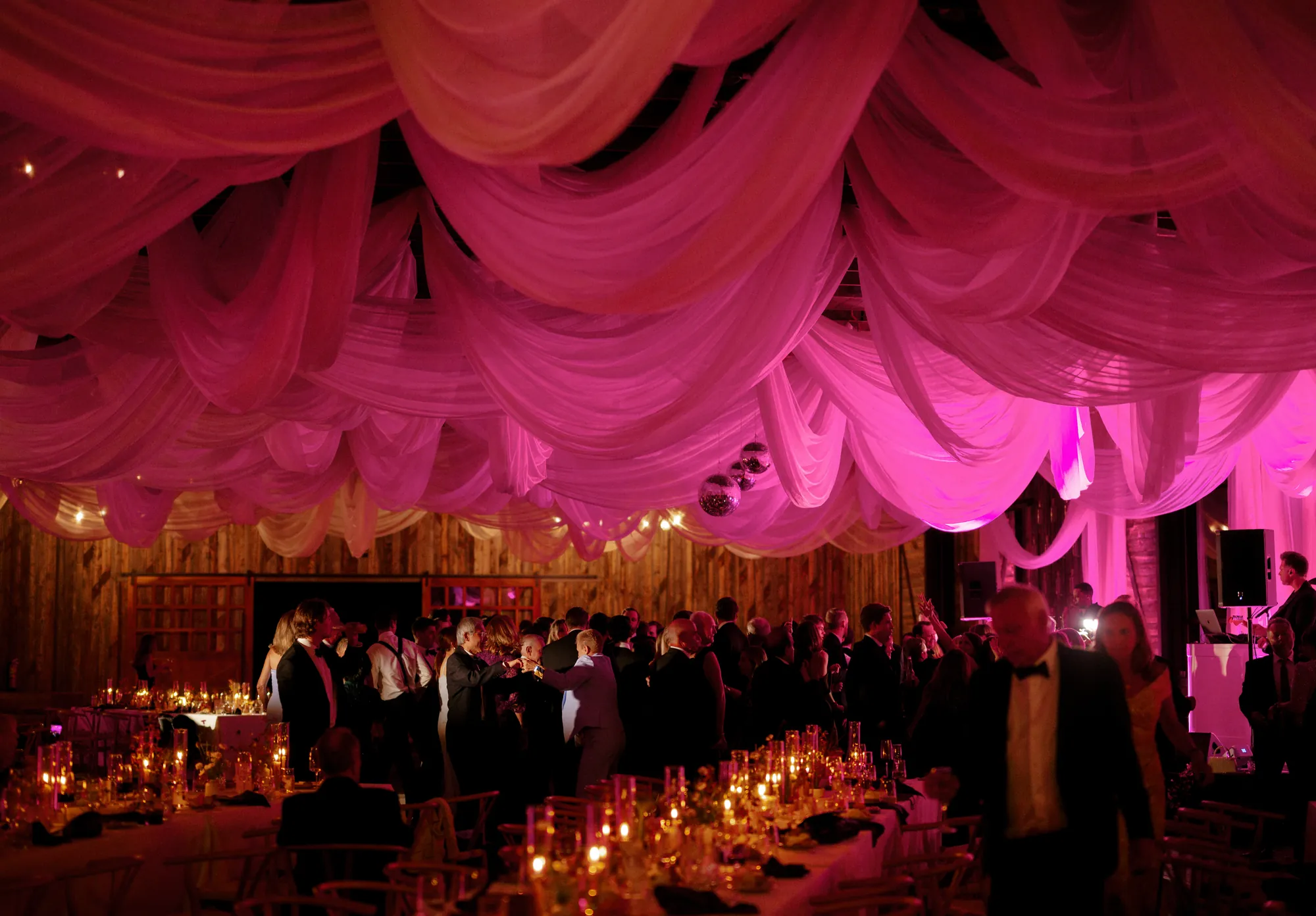 Wedding reception glowing with magenta uplighting beneath dramatic ceiling drapes, guests filling the dance floor